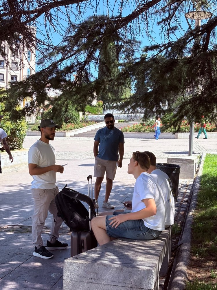 Un estudiante hablando con dos jóvenes en un banco bajo los árboles